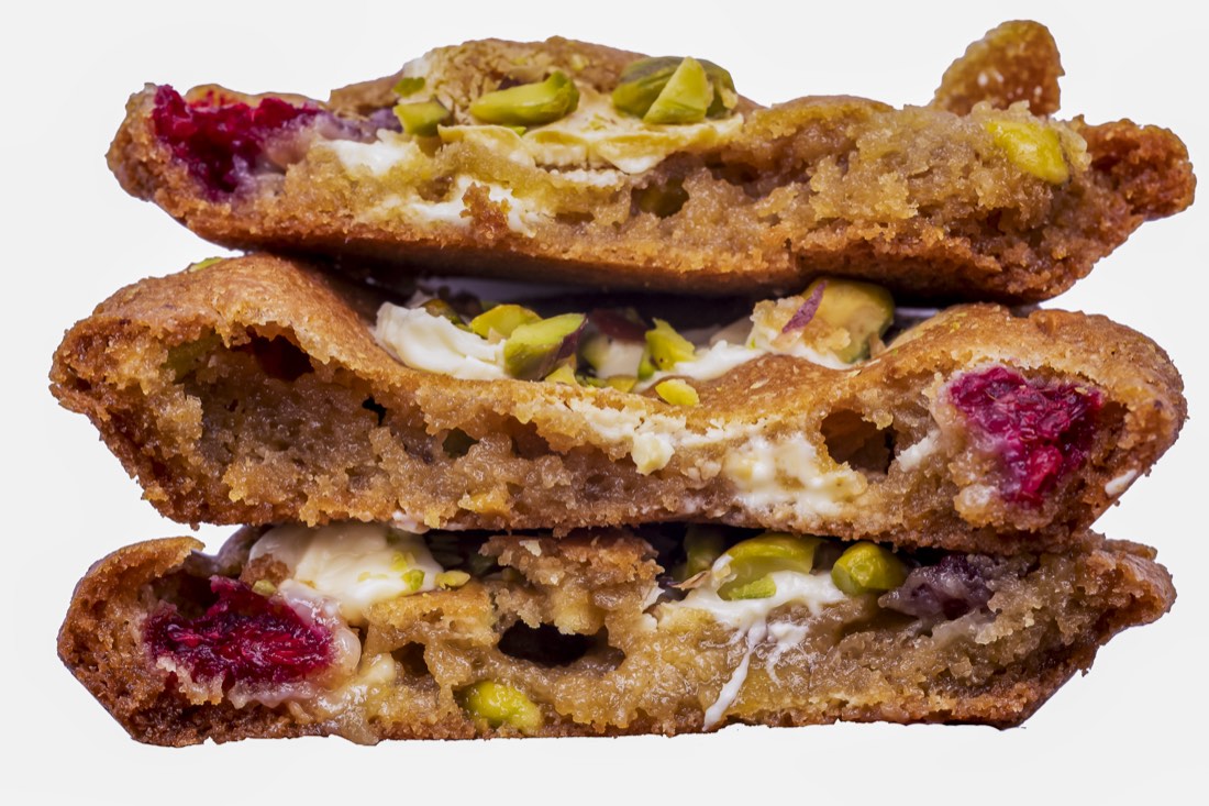 A Raspberry, Pistachio and White Chocolate cookie split open, showing whole raspberries, pistachios and melted white chocolate inside